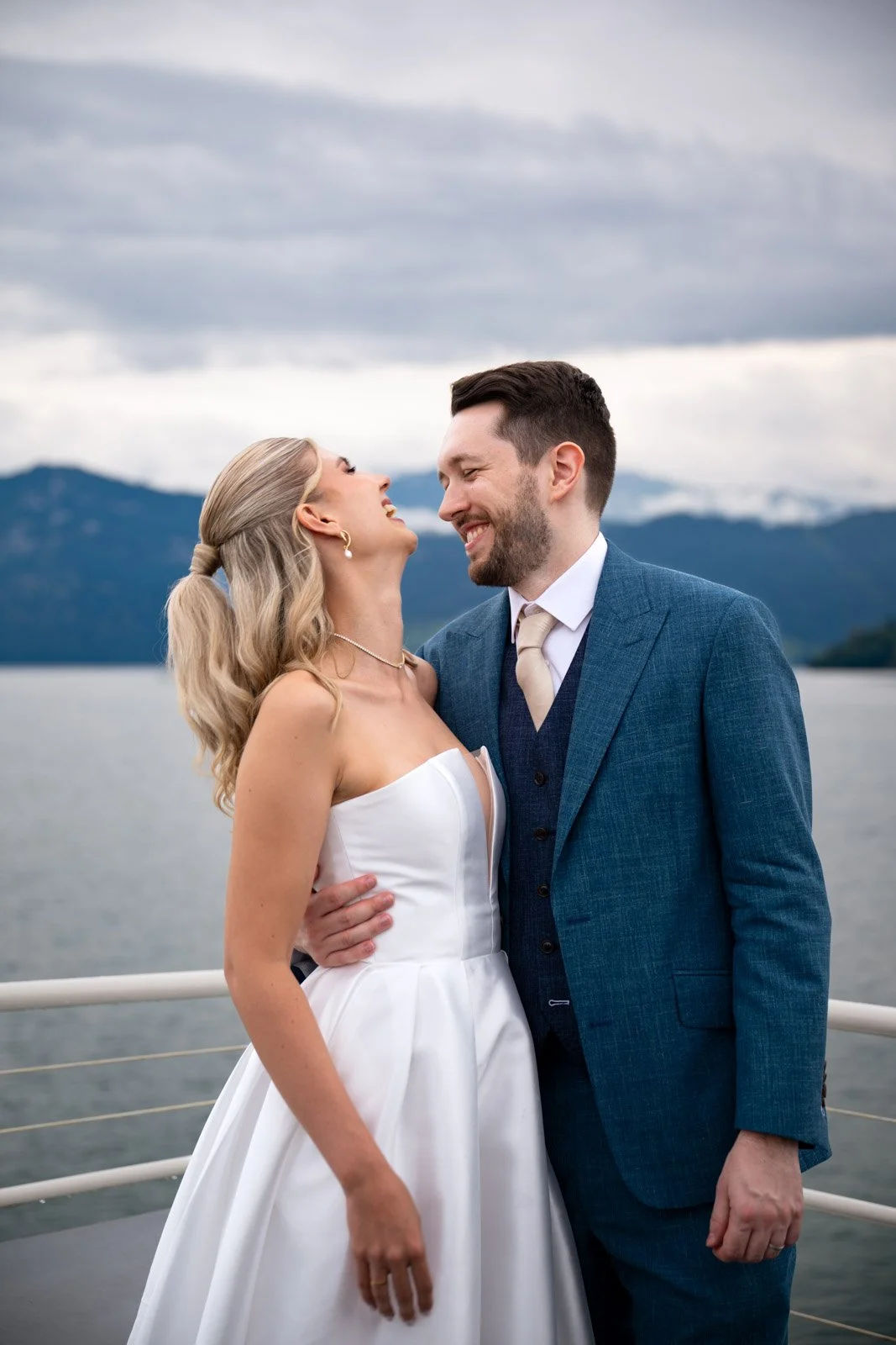 Ein lachendes Brautpaar in formeller Kleidung, die auf einem Boot mit Wasser und Bergen im Hintergrund steht.