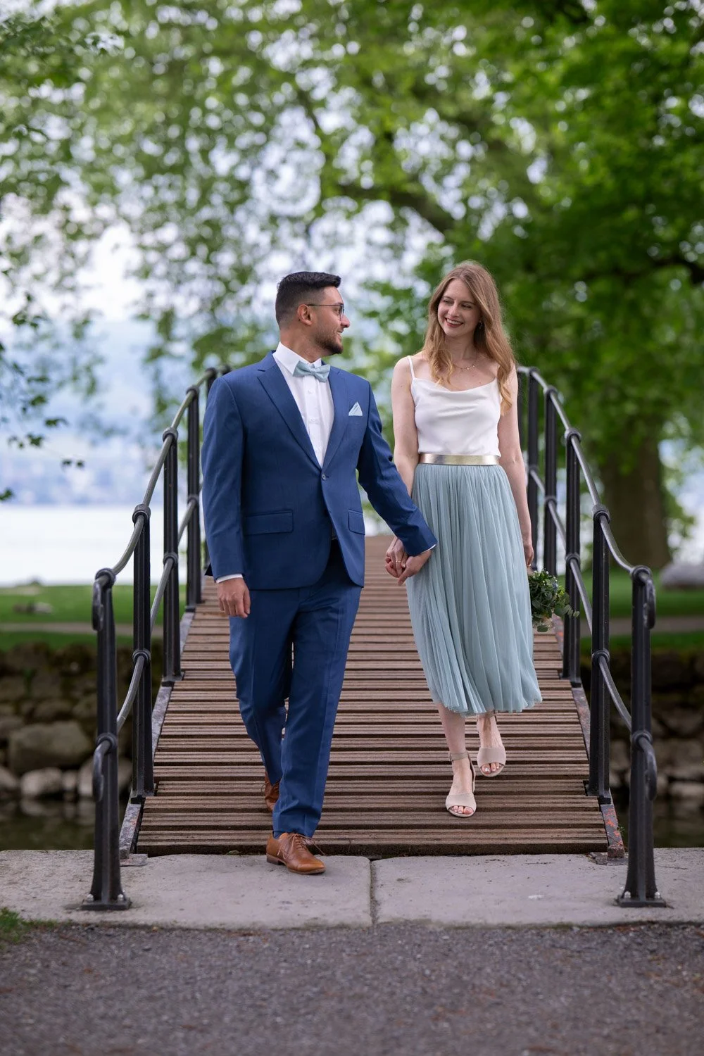 Ein Paar geht Hand in Hand über eine kleine Brücke im Park, umgeben von grünen Bäumen, bei einem festlichen Anlass.