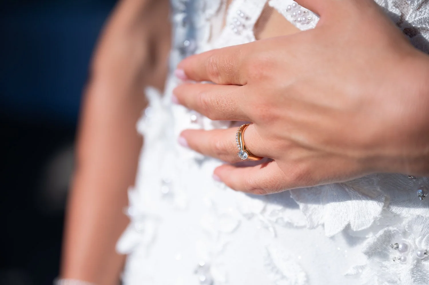 Nahaufnahme einer Hand mit Ehering und Verlobungsring auf einem weißen Brautkleid mit Perlen und Stickereien.