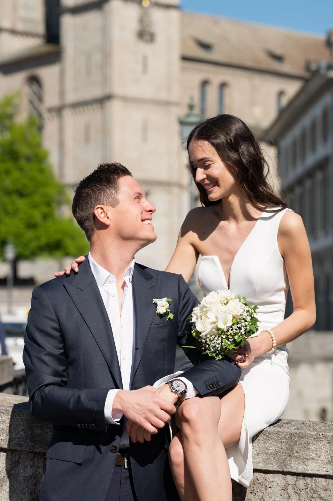 Ein glückliches Paar an ihrer Hochzeit, das draußen vor einer alten Stadtmauer sitzt. Der Mann trägt einen schwarzen Anzug, die Frau ein weißes Hochzeitskleid und hält einen Blumenstrauß in der Hand.