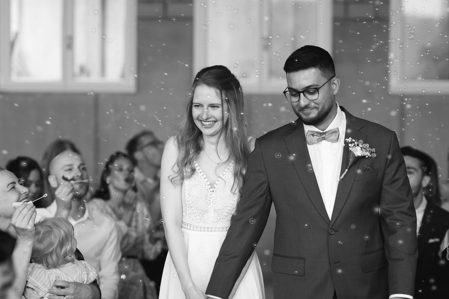 Ein schwarz-weiß Foto eines Brautpaares bei ihrer Hochzeitsfeier, umgeben von jubelnden Gästen, die Seifenblasen machen.