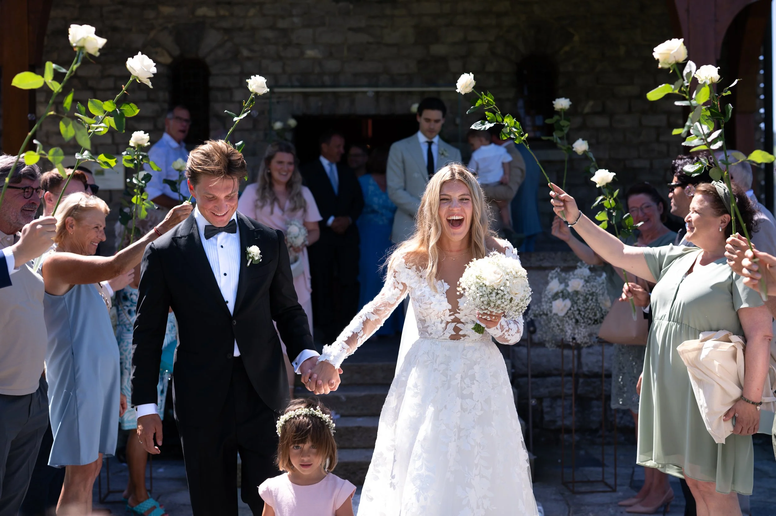 Ein Hochzeitspaar, das sich freut, während sie Hand in Hand gehen, umgeben von Freunden und Familienmitgliedern, die weiße Rosen über ihnen halten, vor einer historischen Steinmauer, bei sonnigem Wetter.