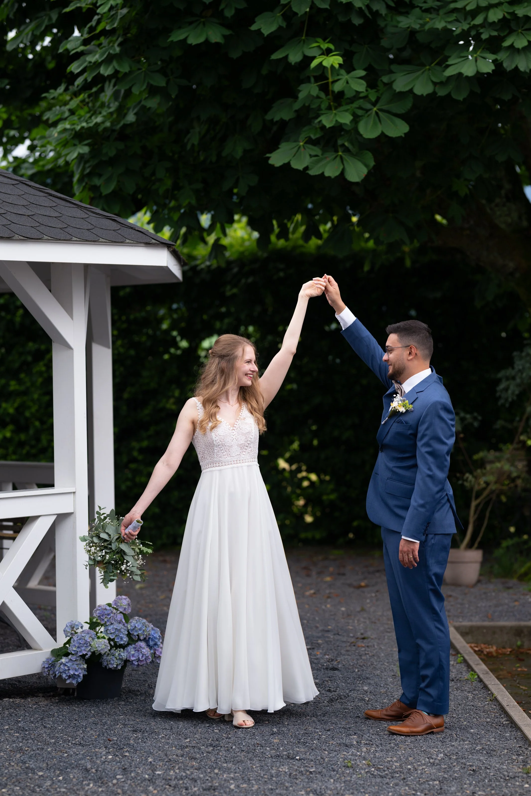 Ein glückliches Brautpaar bei einer Hochzeit im Freien, der Bräutigam hebt die Hand der Braut. Die Braut trägt ein weißes Hochzeitskleid und hält einen Blumenstrauß, während der Bräutigam einen blauen Anzug trägt. Im Hintergrund sind grüne Bäume und 