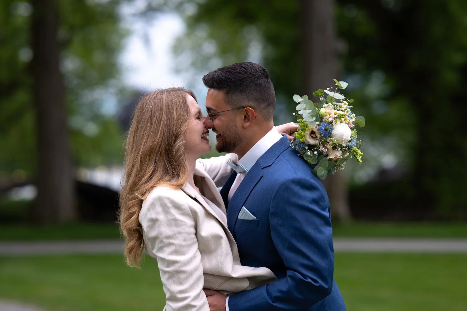 Ein glückliches Paar bei einer Hochzeit, das sich mit den Nasen berührt, im Park mit Bäumen im Hintergrund, der Mann trägt einen blauen Anzug, die Frau ein weißes Jackett, und es gibt einen Blumenstrauß auf der Schulter des Mannes.