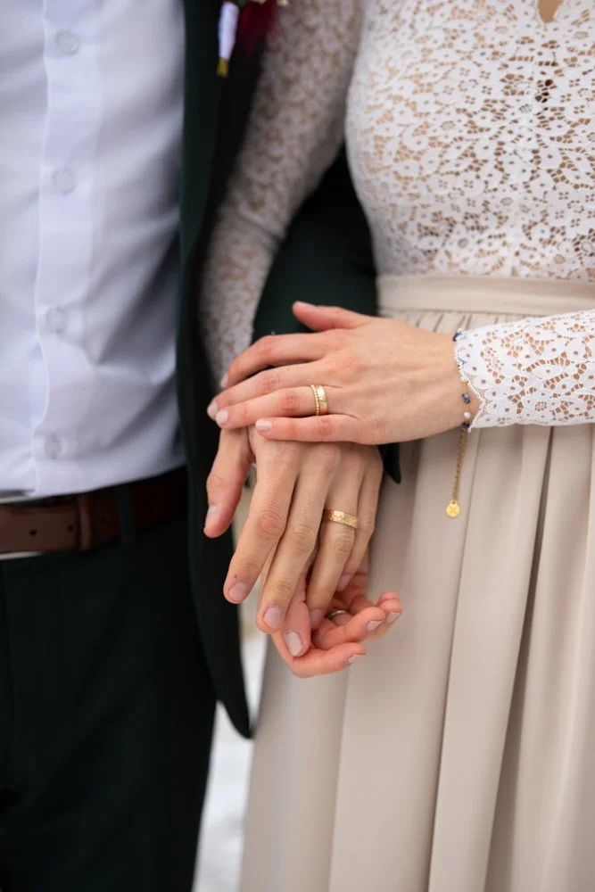 Nahaufnahme von einem Paar, das bei einer Hochzeit ihre Eheringe hält, die Frau trägt ein langärmeliges, weißes Spitzenkleid und der Mann trägt einen schwarzen Anzug.