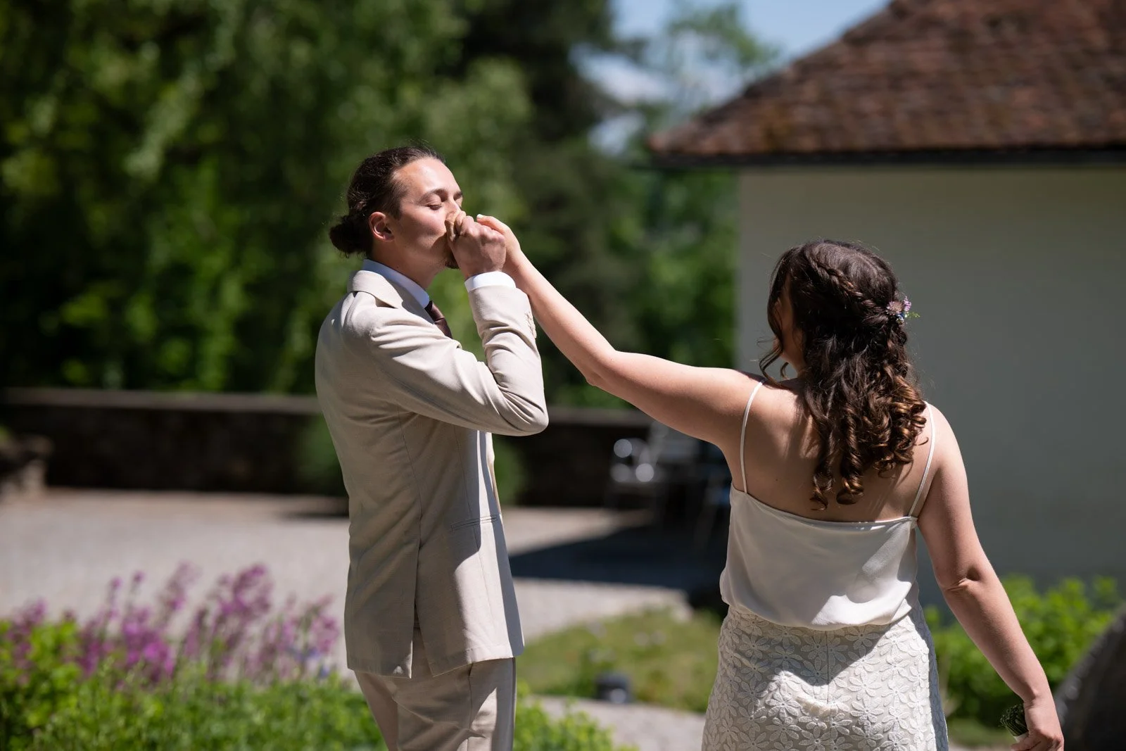 Zwei Frauen, die auf einer Wiese im Sonnenschein stehen. Die eine Frau trägt einen beigen Anzug, die andere ein weißes Kleid. Die Frau im Kleid berührt die Lippen der anderen Frau an der Nase.