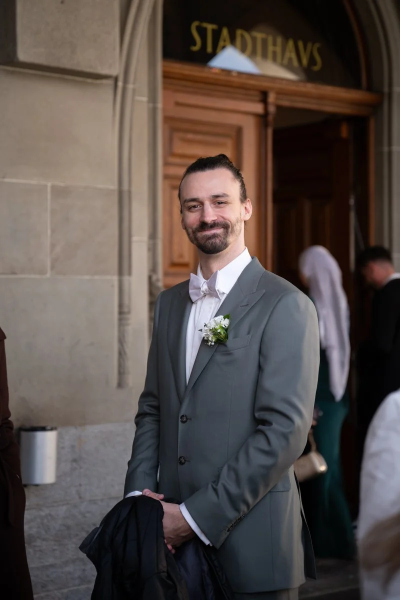 Ein Mann in einem grauen Anzug mit weißem Hemd und Fliege steht vor einer historischen Gebäudefassade. Er trägt ein kleines Blumenportrait am Revers. Im Hintergrund sind weitere Menschen und ein Schild mit dem Wort 'STADTHAUS' zu sehen.