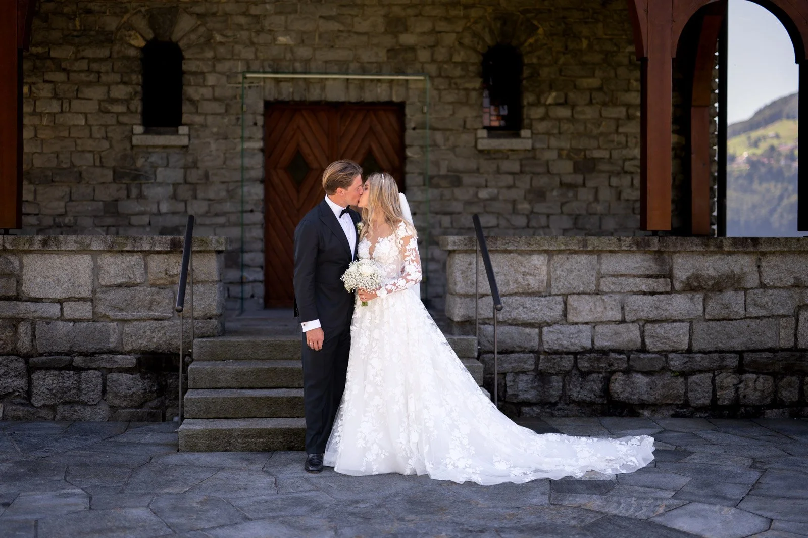 Ein Brautpaar küsst sich vor einer steinernen Kirche. Die Braut trägt ein weißes Hochzeitskleid mit langer Schleppe, der Bräutigam einen schwarzen Anzug. Sie hält einen Blumenstrauß in der Hand.