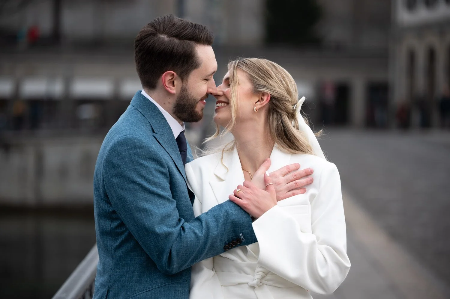 Ein verliebtes Paar, ein Mann in einem blauen Anzug und eine Frau in einem weißen Mantel, stehen sich nahe an einem Fluss in einer Stadt, lächeln und berühren sich zärtlich.