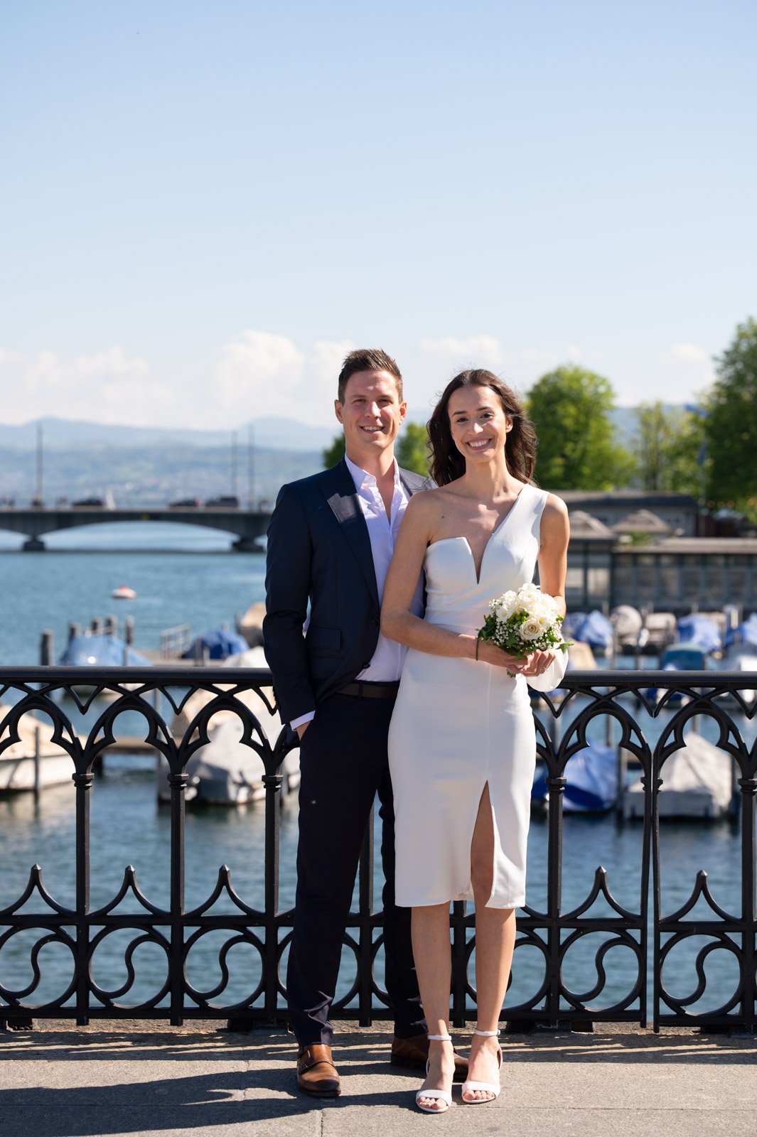 Ein glückliches Paar bei einer Hochzeitsfotografie am Wasser, mit Booten, Brücke und grünen Bäumen im Hintergrund, sonniges Wetter.