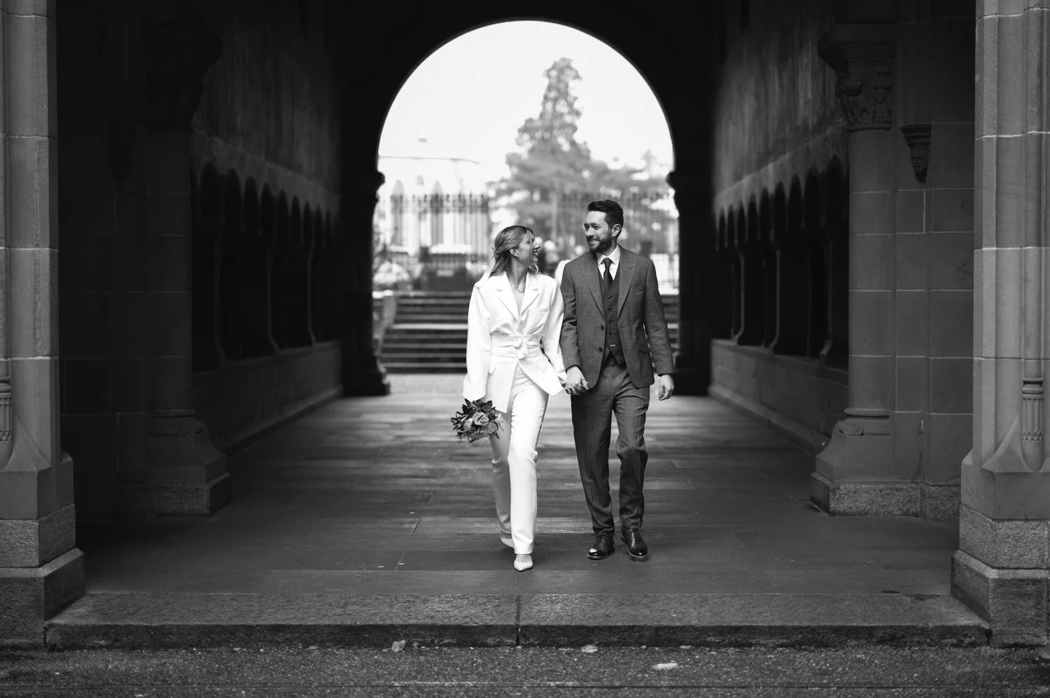 Ein glückliches Paar in formeller Kleidung, das sich Hand in Hand im durch einen Bogen führenden Tunnel auf der Straße beim Spaziergang in einer Stadt bewegt, schwarz-weiße Fotografie.