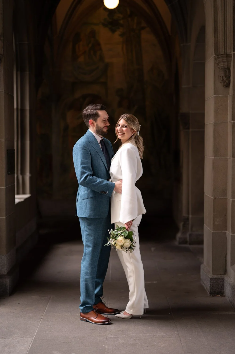 Ein brautpaar in formeller Kleidung, die sich umarmen, im Hintergrund eine Kirche oder Kathedrale mit einem Wandbild. Die Braut hält einen Blumenstrauß, beide lächeln.