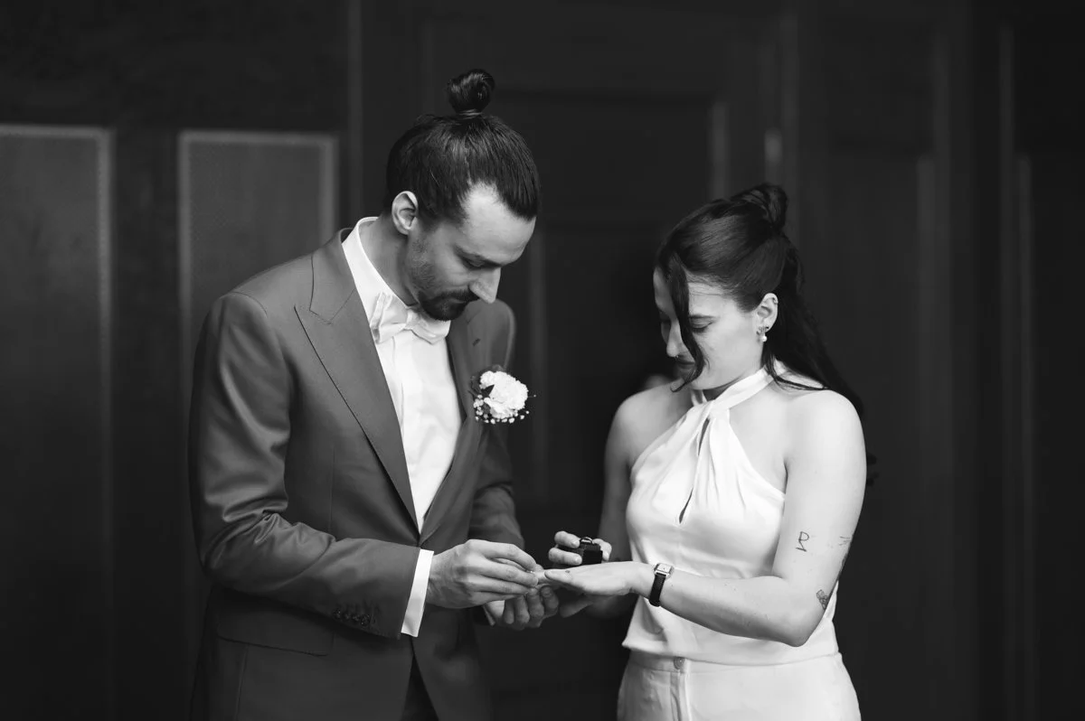 Ein Mann und eine Frau bei einer Zeremonie, vermutlich Verlobung oder Hochzeit, in schwarzer und weißer Kleidung, in einem Indoor-Raum, der Mann steckt ein Schmuckstück an die Frau