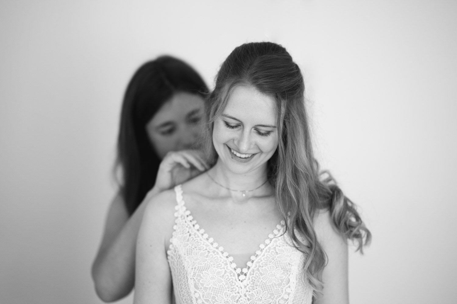 Zwei Frauen, eine schulterfrei gekleidete Frau im Vordergrund, wird von einer zweiten Frau im Hintergrund frisiert, beide lachen und lächeln.