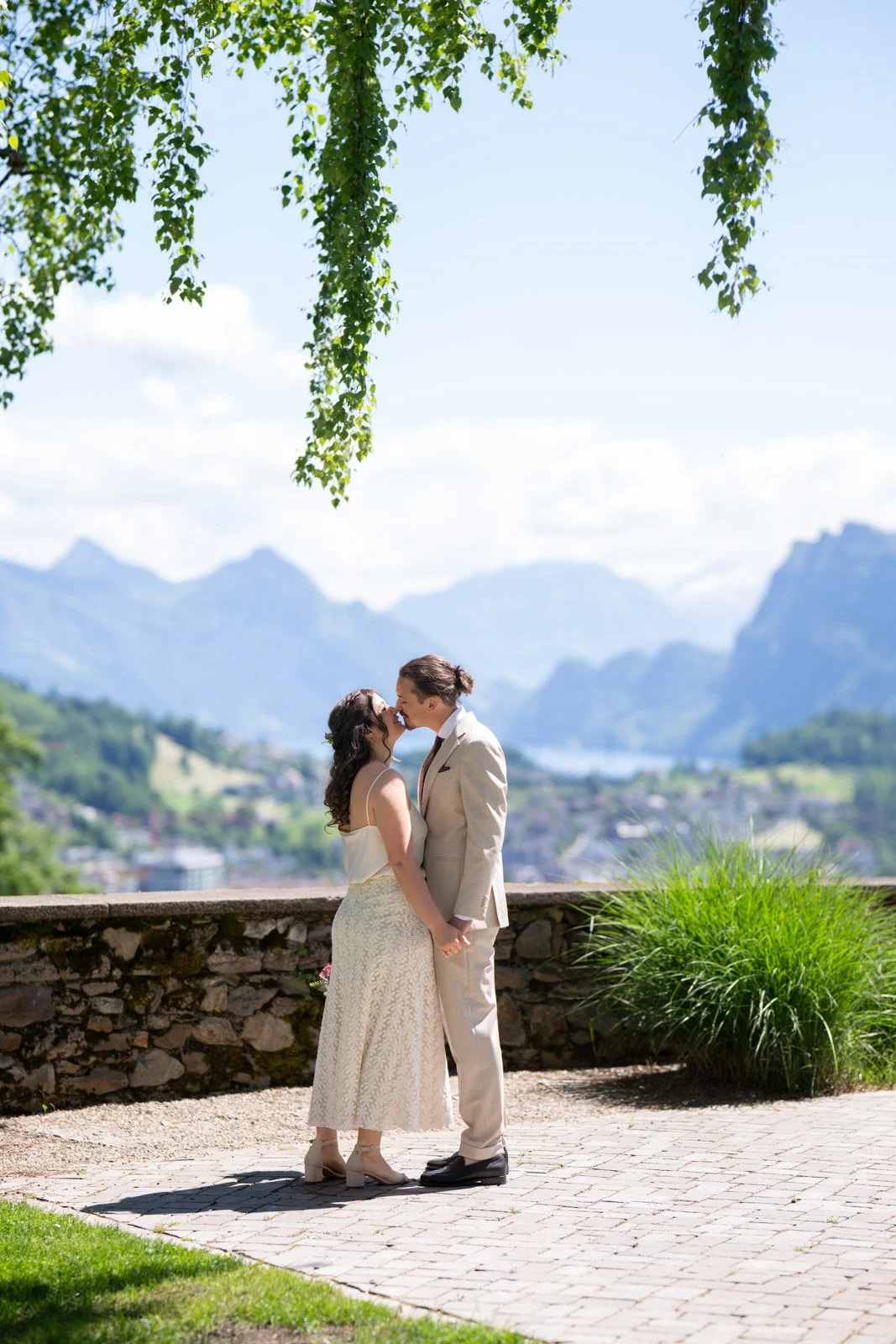 Ein Paar bei einer Hochzeit, das sich küsst, vor einer ländlichen Bergkulisse mit Wasser im Hintergrund, umgeben von Natur, an einem sonnigen Tag.