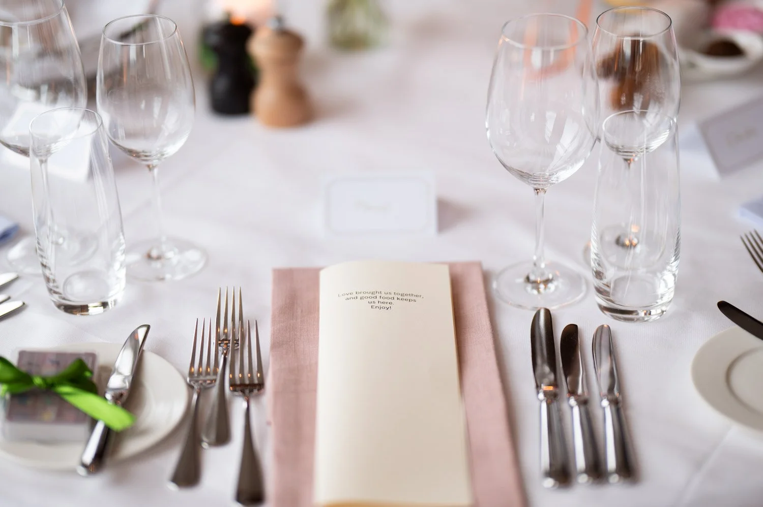 Eleganter gedeckter Tisch mit Weingläsern, Besteck, einem rosa Tuch und einem Menü auf weißem Tischtuch, geeignet für eine formelle Veranstaltung.