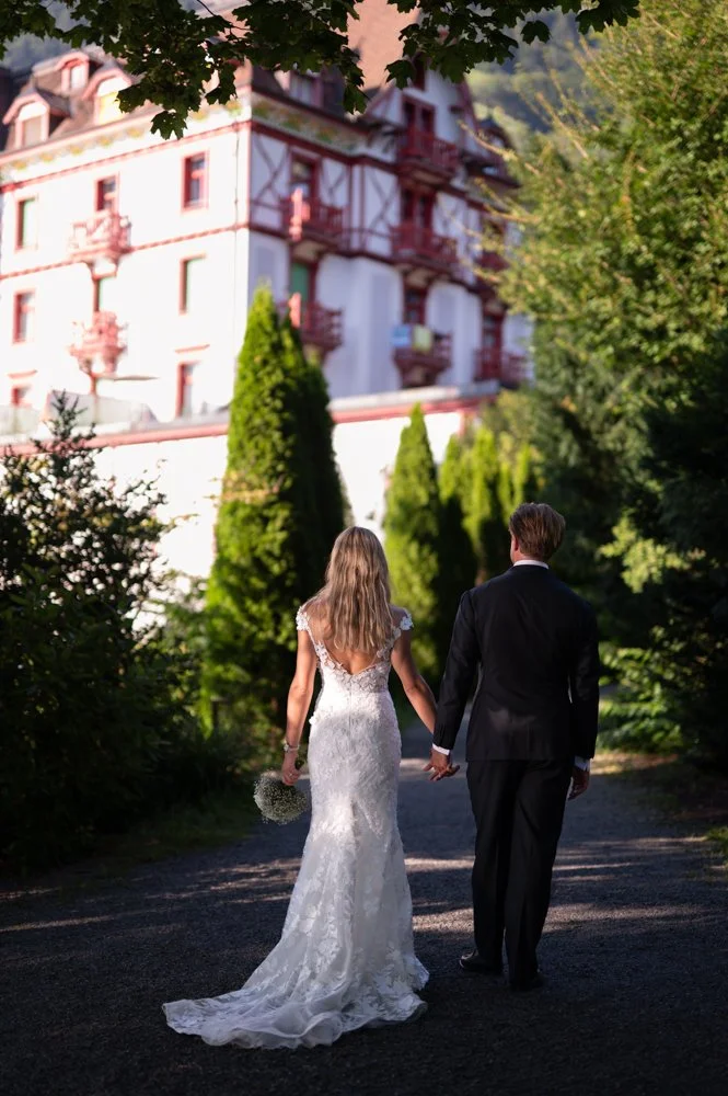 Ein Paar, vermutlich Braut und Bräutigam, hält Händchen und geht auf einem Pfad im Grünen, mit einem großen Haus im Hintergrund.