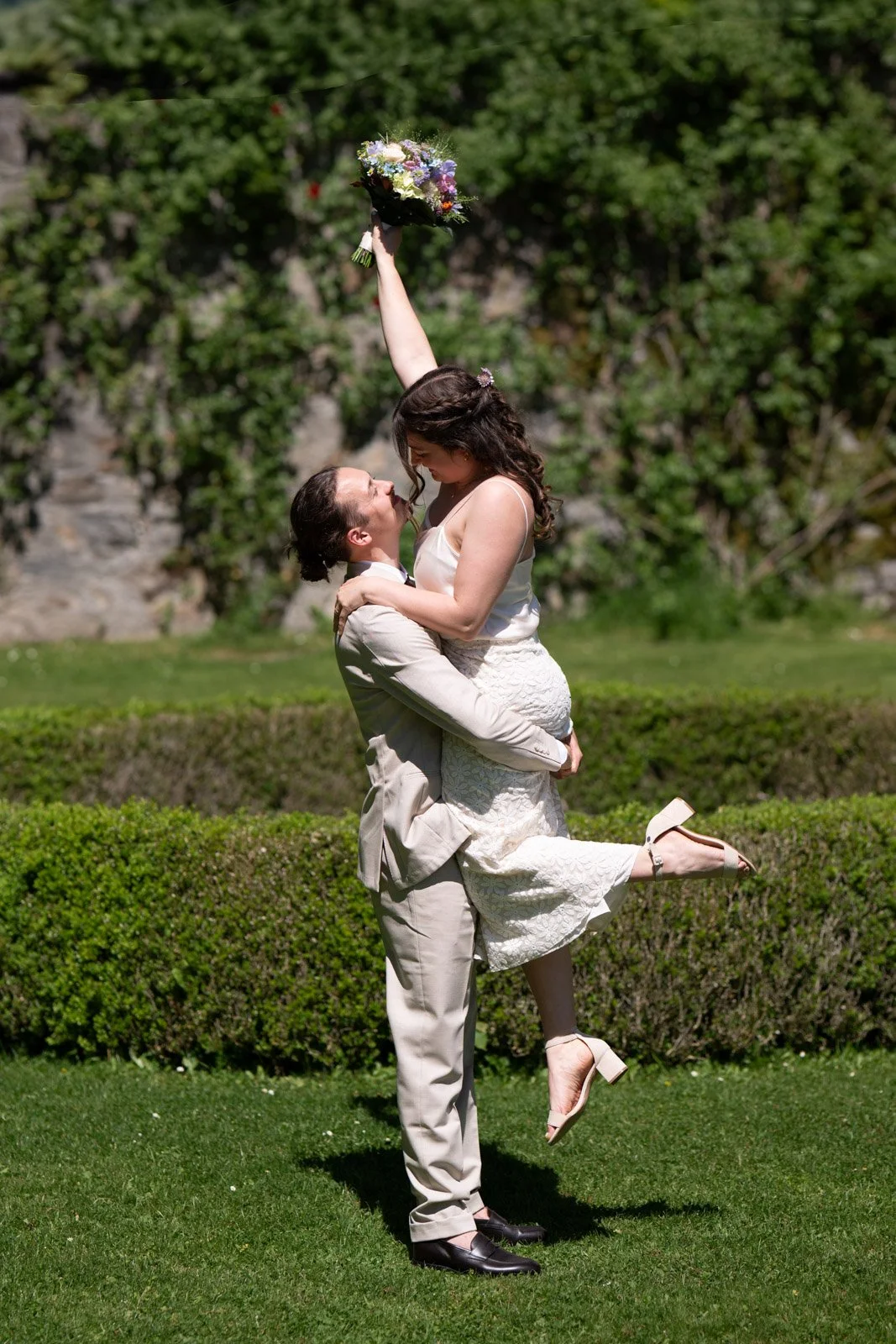 Ein glückliches Paar bei einer Hochzeit im Freien. Der Mann hebt die Frau hoch, während sie eine Blumenstrauß hält. Es ist sonnig und grün.
