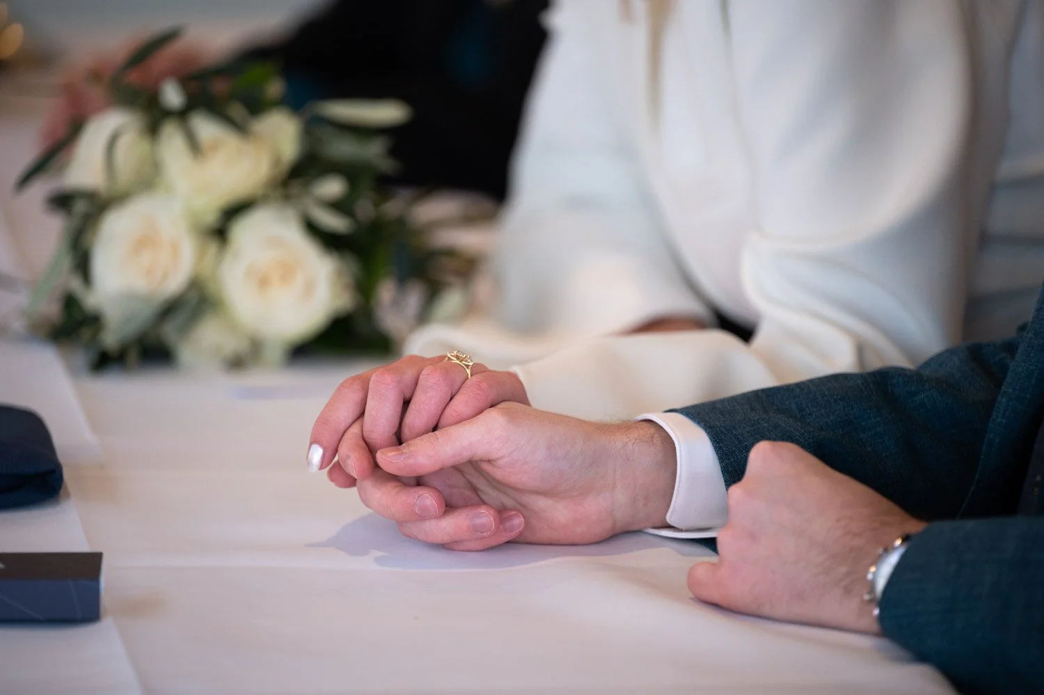 Ein Brautpaar hält bei einer Trauzeremonie ihre Hände auf einem Tisch, im Hintergrund eine Blumenstrauß. 