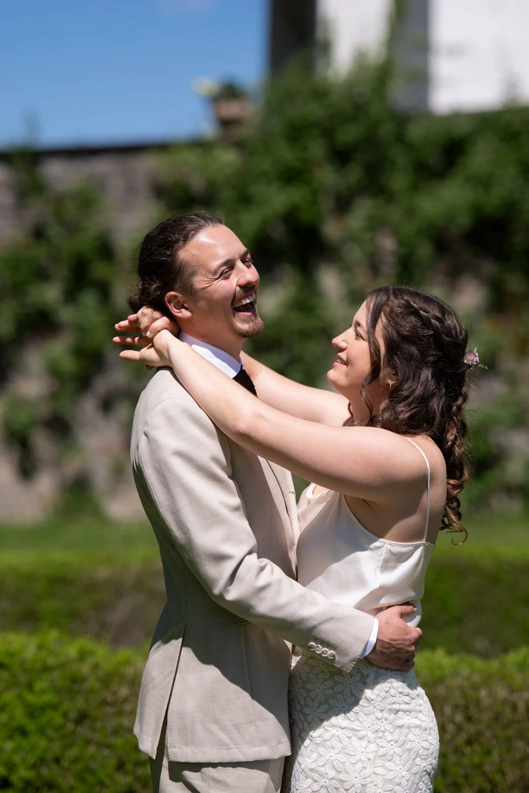 Ein glückliches Paar bei einer Hochzeit im Freien, umarmt sich und lacht im Sonnenlicht, im Hintergrund grüne Bäume.