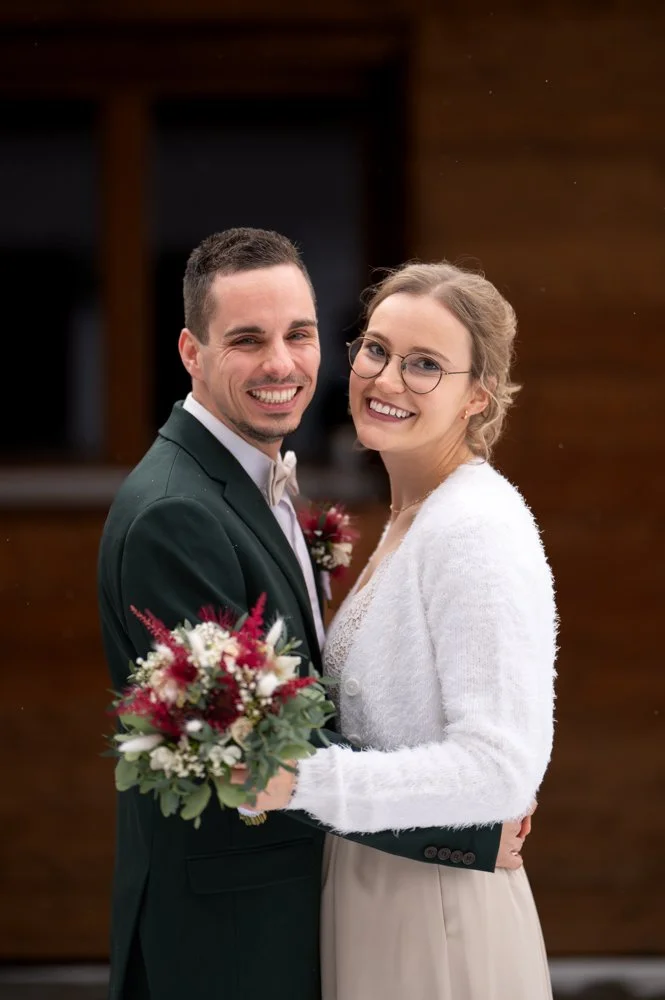 Ein lachendes Paar bei einer Hochzeit, der Mann trägt einen schwarzen Anzug mit Fliege und die Frau trägt ein weißes Kleid mit Brille, hält einen Blumenstrauß, vor einem Holzhintergrund.