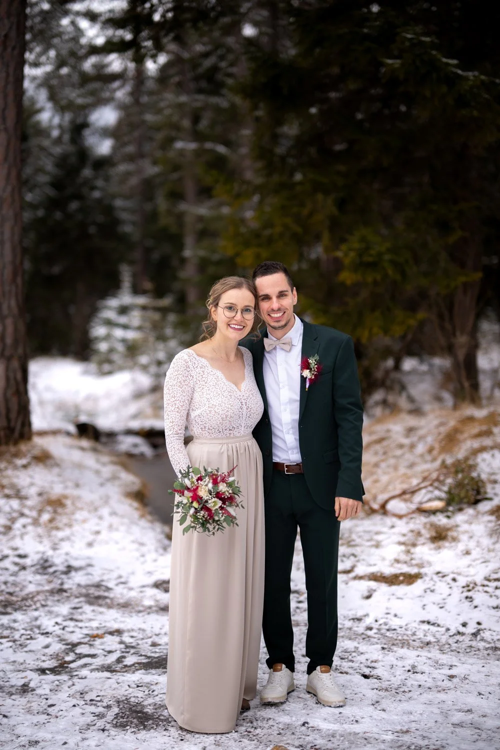 Ein Heiratspaar in der Schneelandschaft, lachend, umgeben von Tannenbäumen, Frau trägt ein langes, weißes Kleid, Mann trägt ein schwarzes Anzug mit Fliege, beide halten Blumen und wirken glücklich.