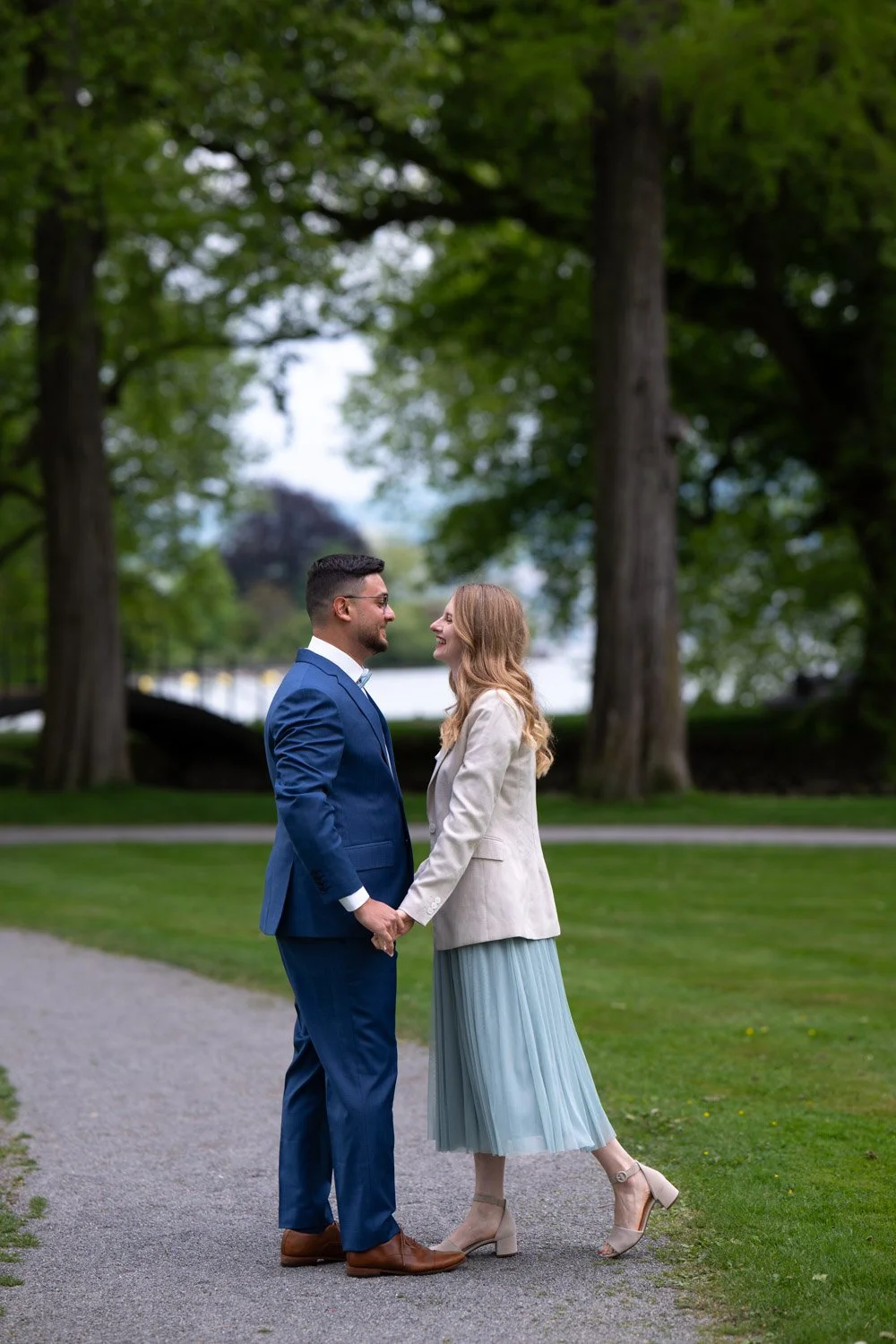 Ein Mann und eine Frau stehen sich in einem Park gegenüber, halten sich an den Händen und lächeln sich an, umgeben von großen Bäumen und grünem Gras.