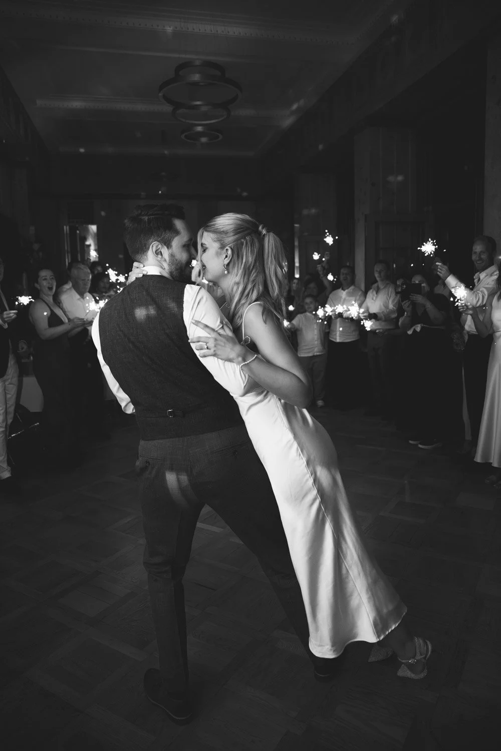 Ein Paar tanzt in einer Hochzeitsfeier, umgeben von Gästen, die Leuchtstäbe halten, in einem festlichen Raum.