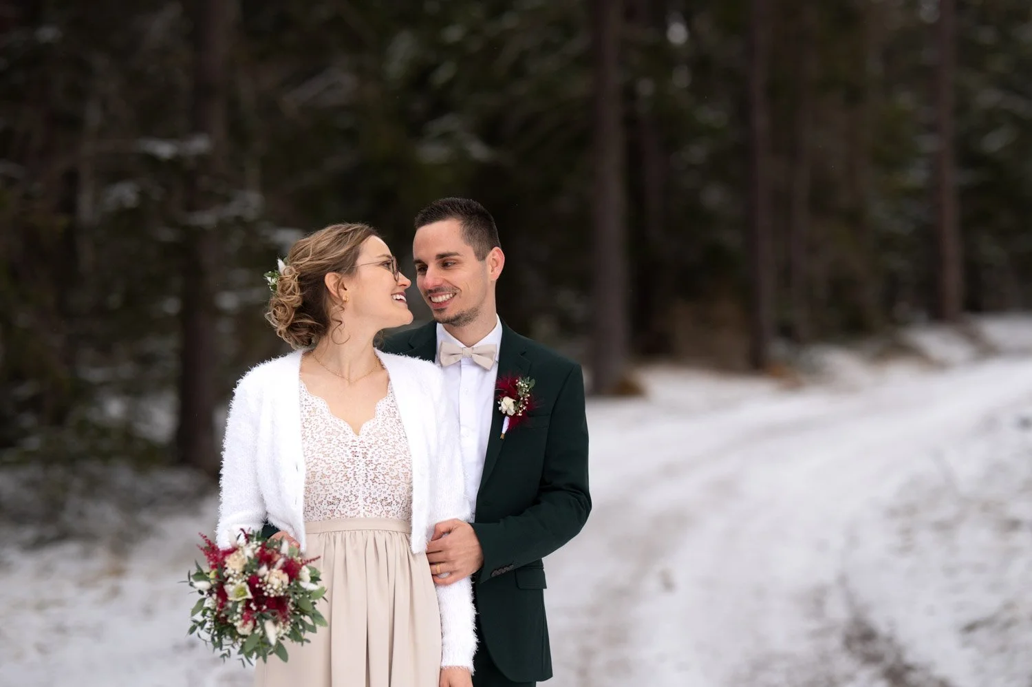 Ein glückliches Brautpaar im Schnee in einem Wald, die Frau hält einen Blumenstrauß, beide blicken sich liebevoll an.