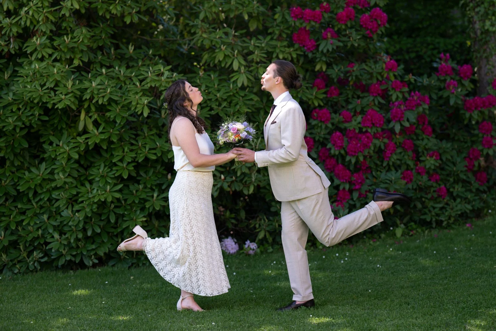 Ein Paar in formeller Kleidung, der Mann in einem hellen Anzug und die Frau in einem weißen Kleid, stehen auf einem grünen Rasen vor einem Busch mit rosa Blumen. Sie stehen sich gegenüber, halten eine Blumenstrauß, und der Mann ist auf einem Bein, wä