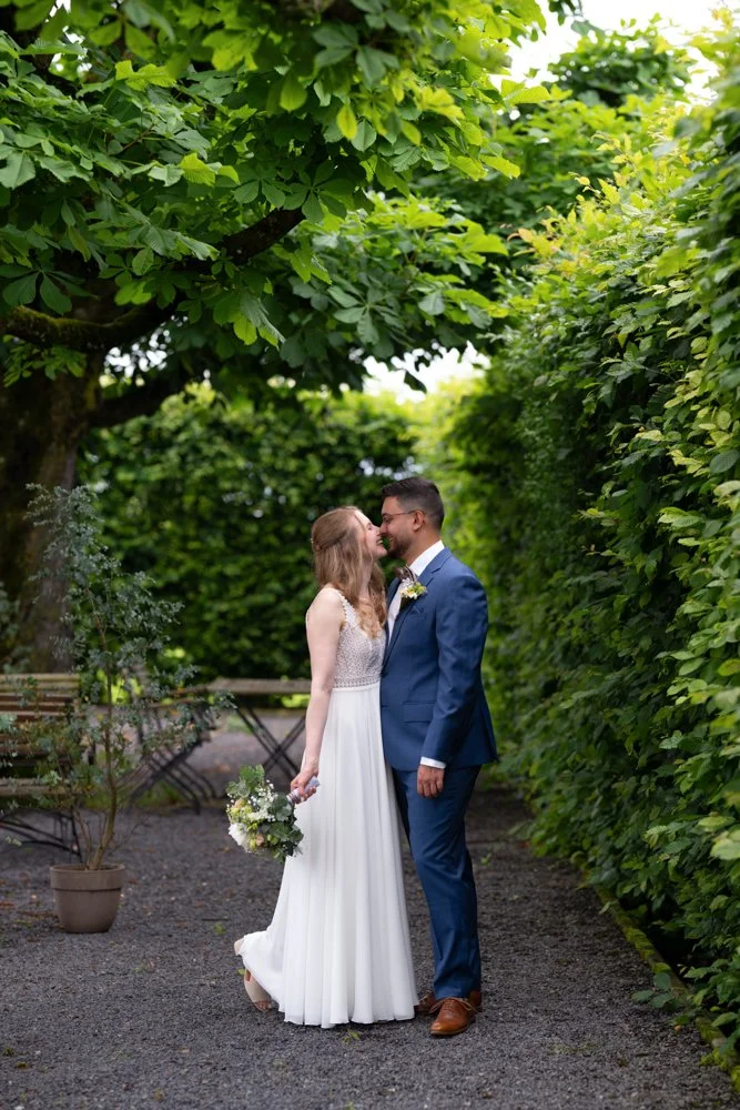 Ein verliebtes Hochzeitspaar steht in einem grünen Garten, umgeben von Büschen und Bäumen, die sich an einem verschlungenen Weg befinden. Die Frau trägt ein weißes Hochzeitskleid und hält einen Blumenstrauß, während der Mann einen blauen Anzug trägt.