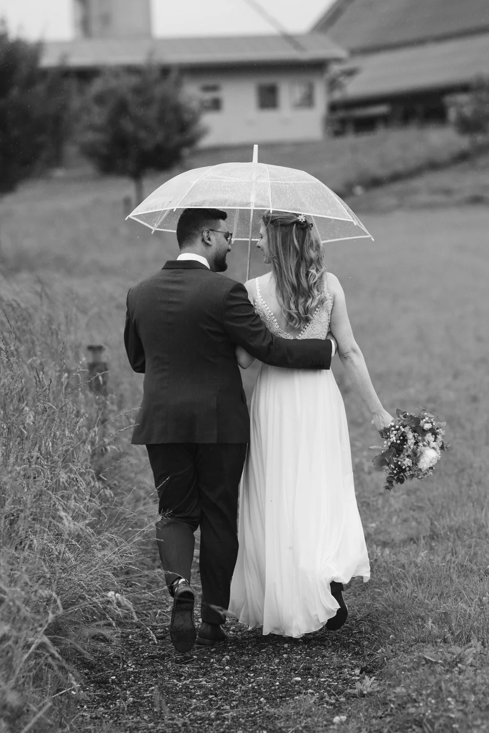 Ein Brautpaar läuft Hand in Hand im Regen, unter einem transparenten Regenschirm, die Braut hält einen Blumenstrauß in der Hand, beide lachen und schauen sich liebevoll an, im Hintergrund Häuser und Bäume, in Schwarz-Weiß fotografiert.