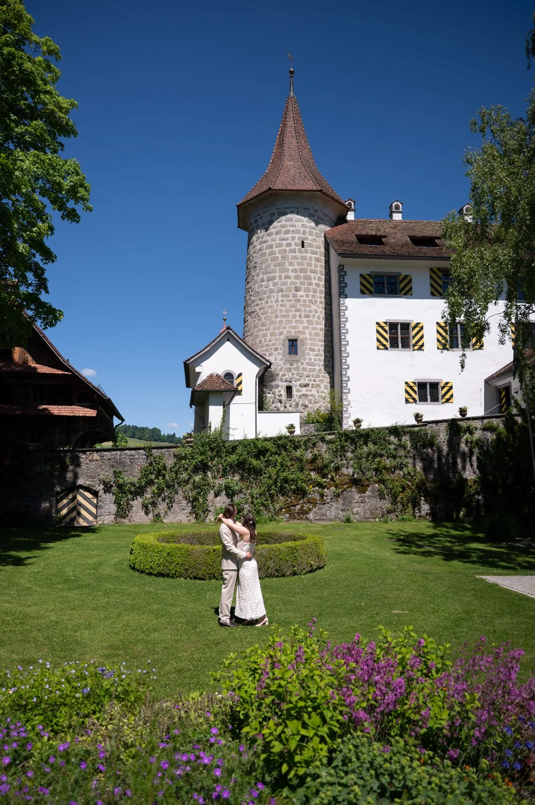 Ein Brautpaar tanzt auf einem gepflegten Rasen vor einer alten Burg im Sonnenschein. Im Vordergrund blühen farbige Blumen, im Hintergrund ist eine Burg mit runden Türmen und weißen Wänden zu sehen, umgeben von Bäumen und einer Mauer.