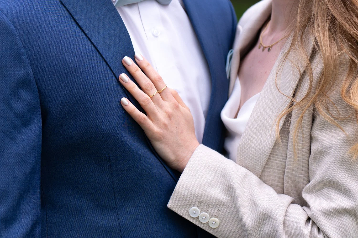 Nahaufnahme eines Paares, das sich nahe steht. Die Frau hat die Hand auf die Brust des Mannes gelegt, beide tragen elegante Kleidung, der Mann in einem blauen Anzug und die Frau in einem cremefarbenen Blazer.
