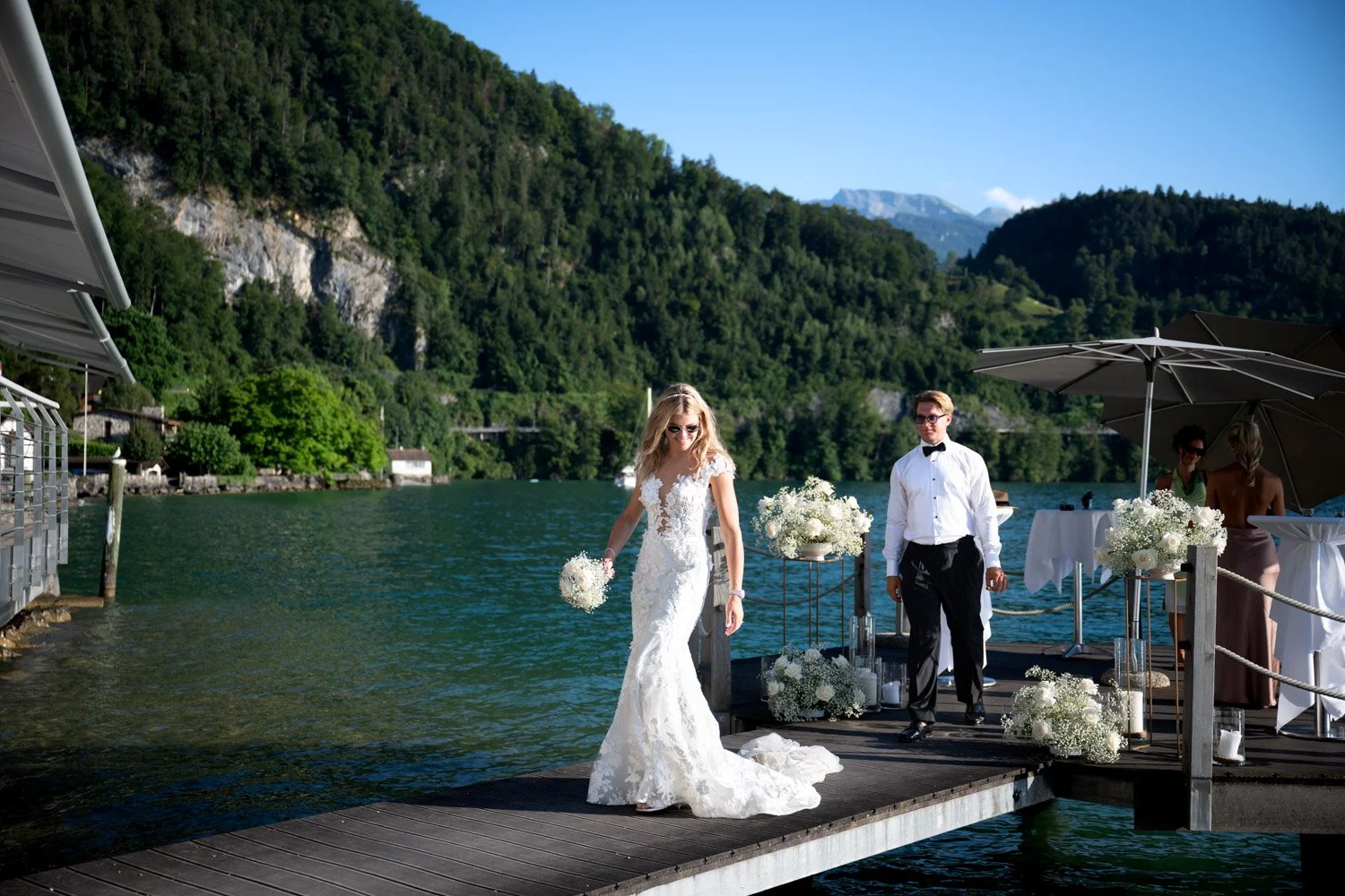 Hochzeit am See mit Braut in weißem Kleid, Bräutigam in Smoking, im Hintergrund Berge und Wasser, sonniger Tag.