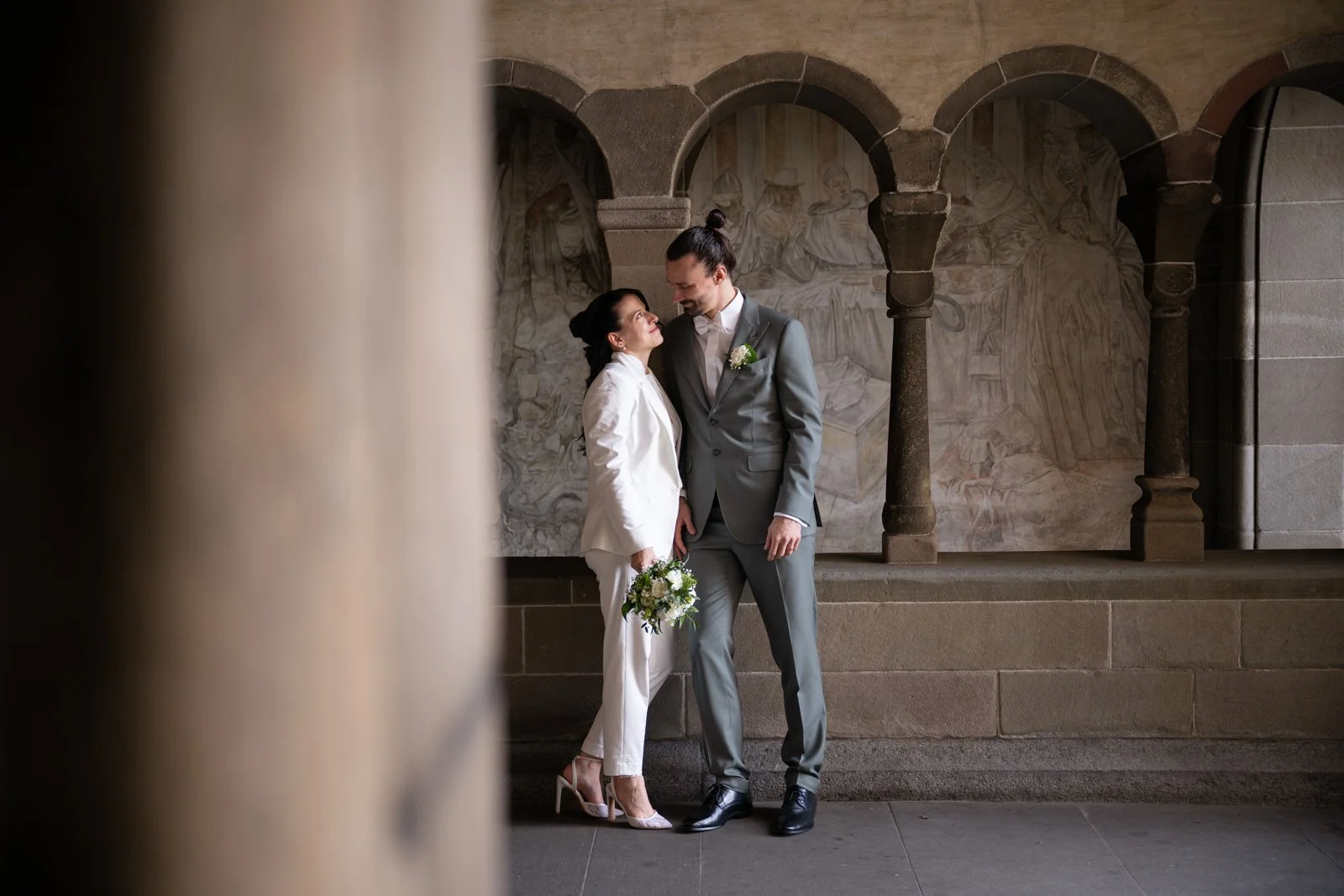 Ein Brautpaar steht in einer historischen, steinernen Kulisse. Die Braut trägt einen weißen Anzug mit hohen Schuhen und hält einen Blumenstrauß, während der Bräutigam einen grauen Anzug mit weißen Hemd und dunklen Schuhen trägt.