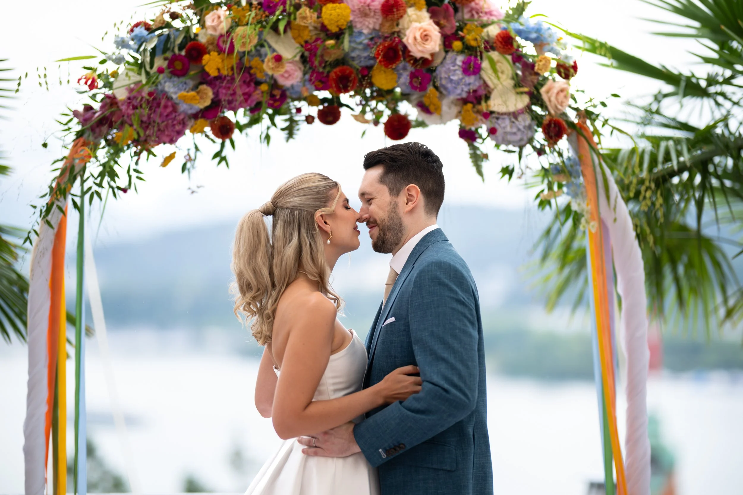 Ein heiratswilliges Paar steht sich bei ihrer Hochzeit unter einem Blumenbogen aus bunten Blumen und Palmen im Hintergrund gegenüber.