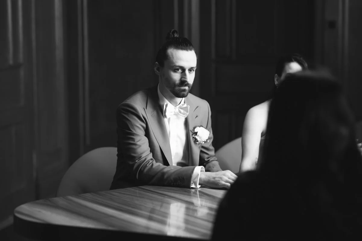 Schwarz-weiß-Foto eines Mannes in einem Anzug mit Hemd und Fliege, sitzend an einem runden Tisch, mit einem Blumenstrauß am Revers, in einem Raum mit dunklen Holzwänden.