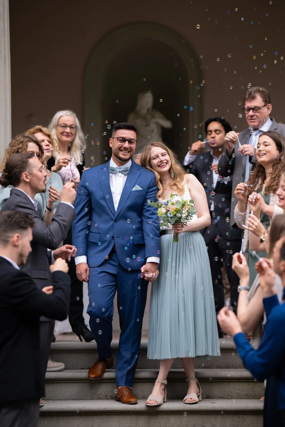 Ein Brautpaar, das auf den Treppen einer Kirche steht, umgeben von Gästen, die Seifenblasen machen, bei einer Hochzeit.