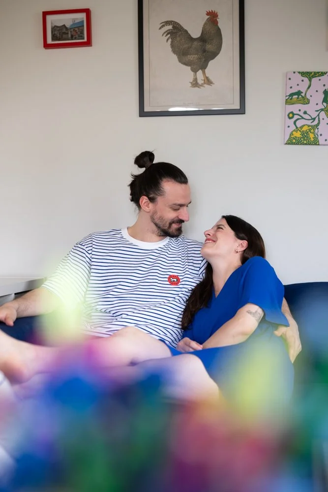 Ein Mann und eine Frau sitzen auf einem Sofa, sie schauen sich liebevoll an. Im Hintergrund hängen Bilder an der weißen Wand, darunter ein Bild eines Huhns.