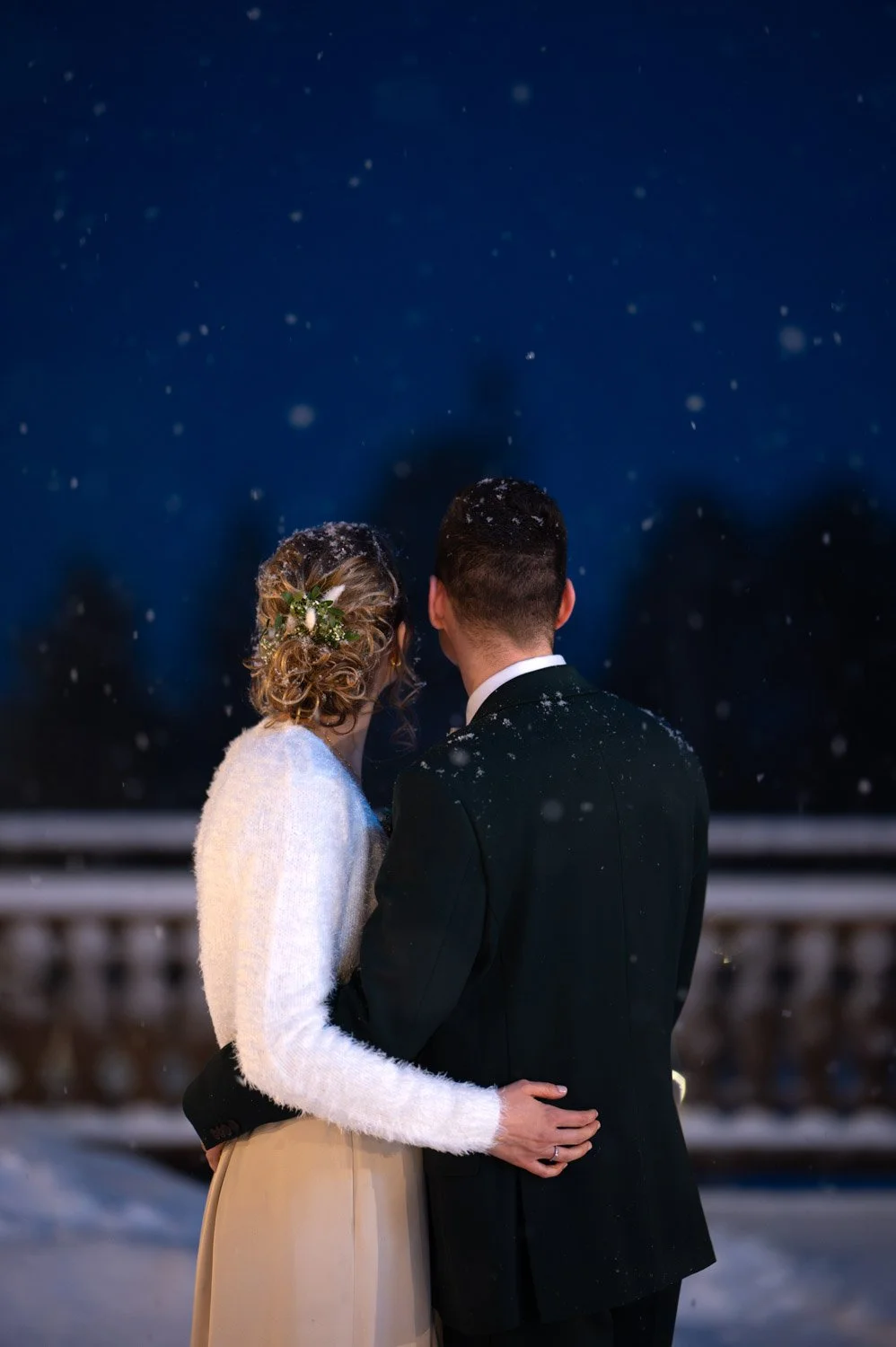 Ein Paar, das bei Nacht im Schnee steht, sieht sich liebevoll an, mit Schneeflocken auf ihren Haaren und Kleidung.