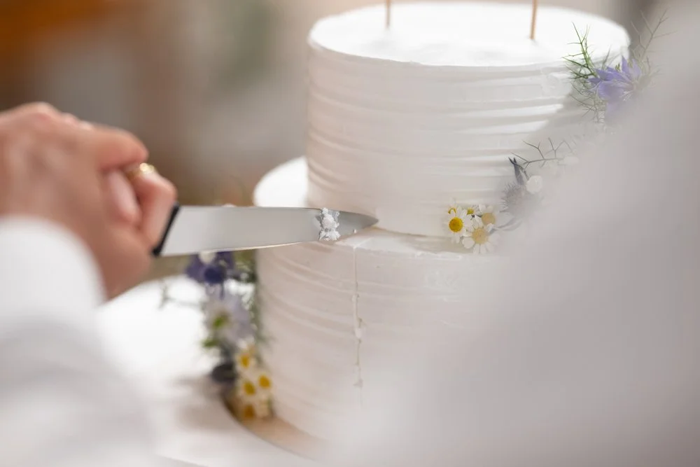 Eine Person schneidet eine weiße Hochzeitstorte mit einem Messer, die Torte ist verziert mit kleinen Blumen.