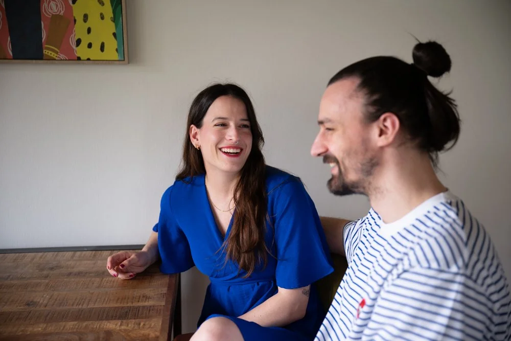 Ein Mann und eine Frau sitzen an einem Holztisch, lachen und unterhalten sich in einem Raum mit weißer Wand. Im Hintergrund ist ein farbenfrohes Kunstwerk an der Wand zu sehen.