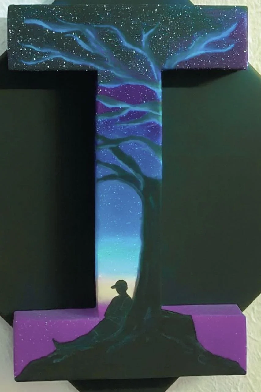 Hand-painted cardboard piece on canvas in the shape of a capital I depicting a night sky with stars, a large tree with twisting branches, and a silhouette of a person sitting by the base of the tree.