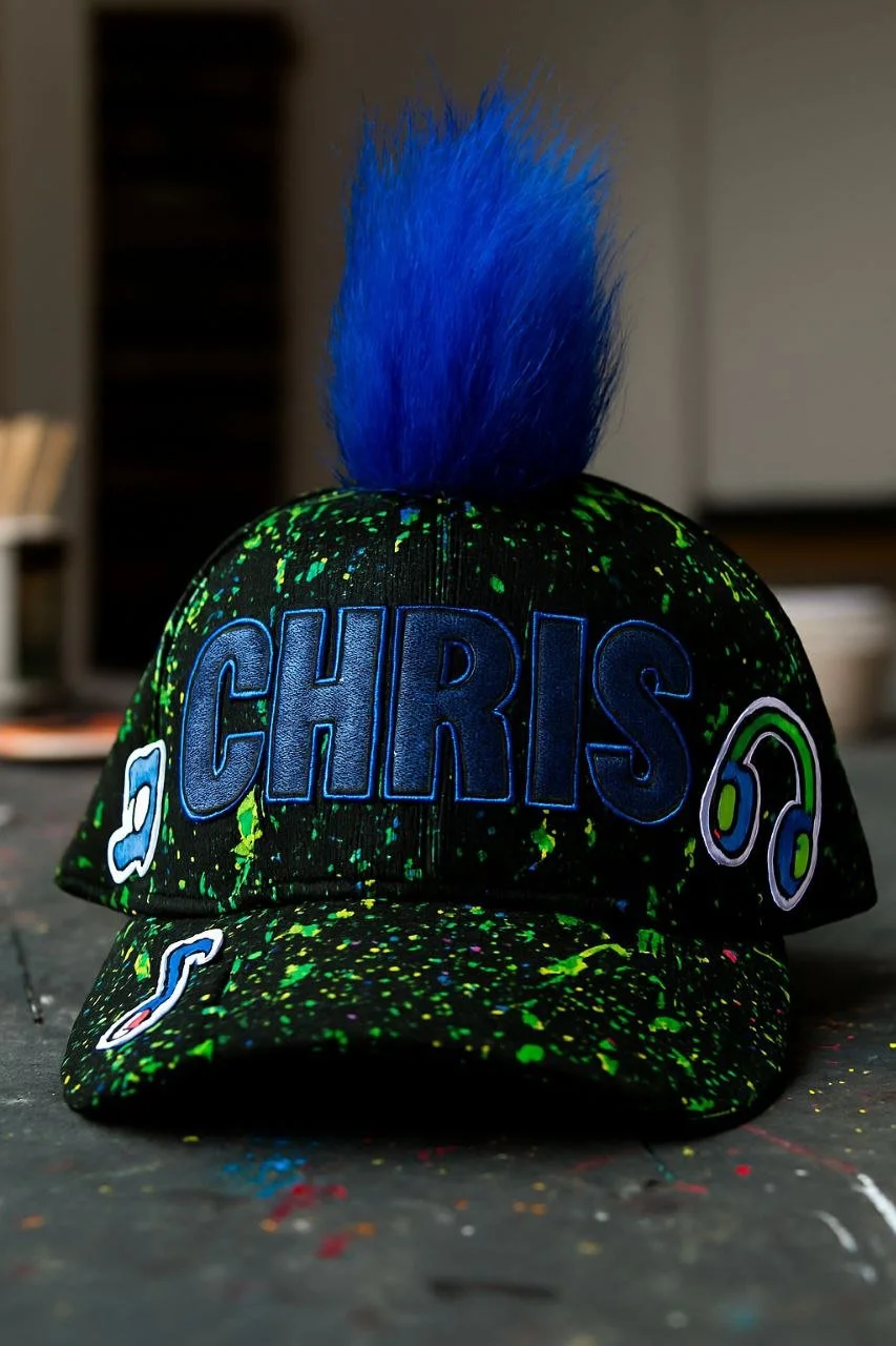 Black cap with neon green splatters, featuring large blue embroidered letters spelling 'CHRIS', decorated with cartoon-like white and blue headphones and musical notes, topped with a large blue pom-pom.