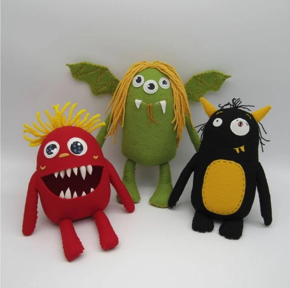 Three handmade plush monster toys, one red with yellow eyes and sharp teeth, one green with three eyes and yellow yarn hair, and one black with horns and a yellow belly, posed against a plain white background.