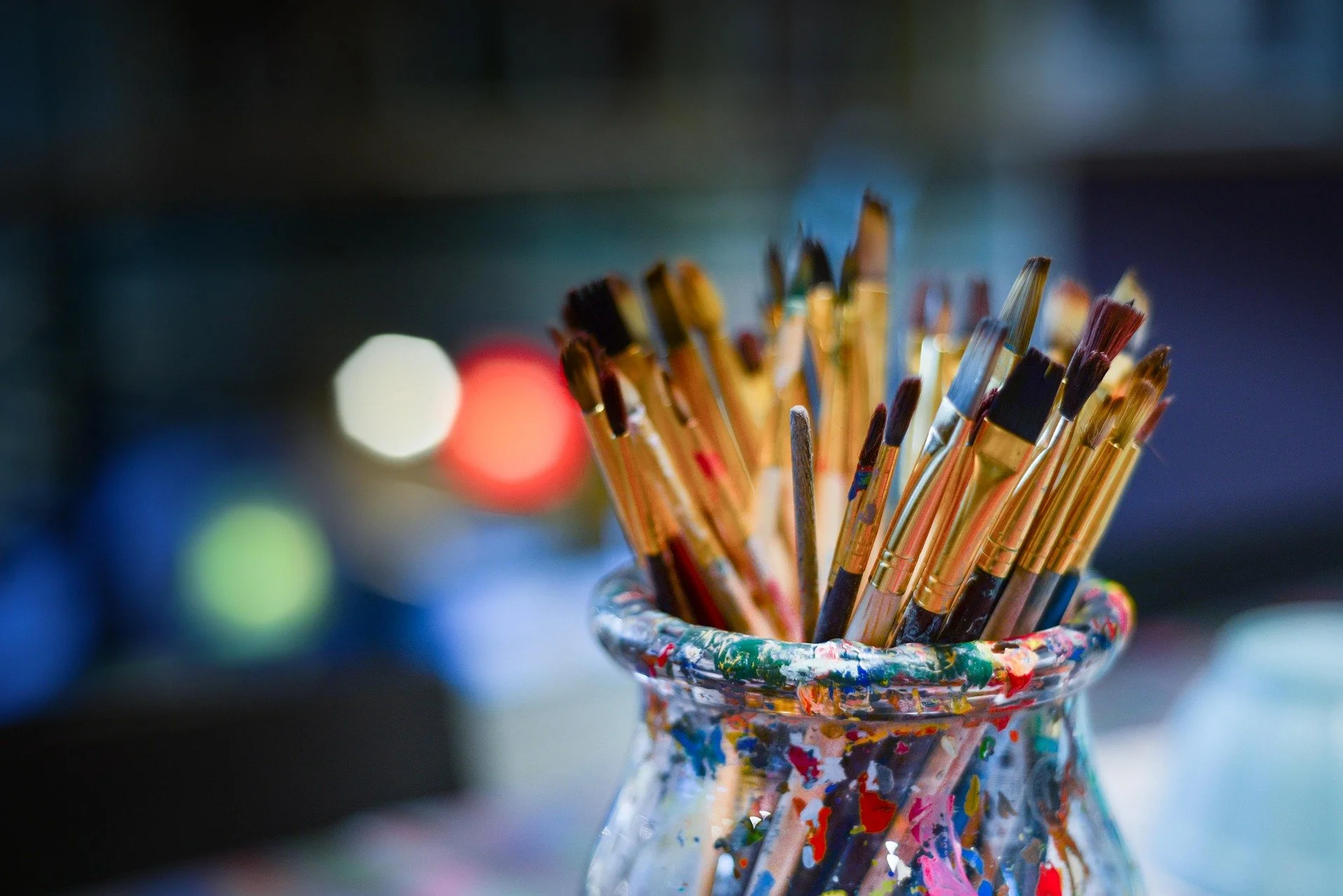 A glass jar filled with paintbrushes, some with paint on the handles, sitting on a surface with a bokeh background.