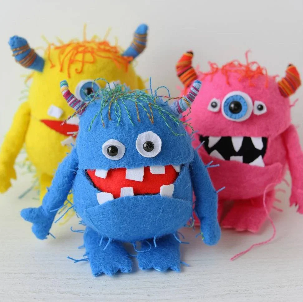 Colorful plush monster toys with striped horns, googly eyes, big mouths, and fuzzy wool yarn hair, posed on a white surface.