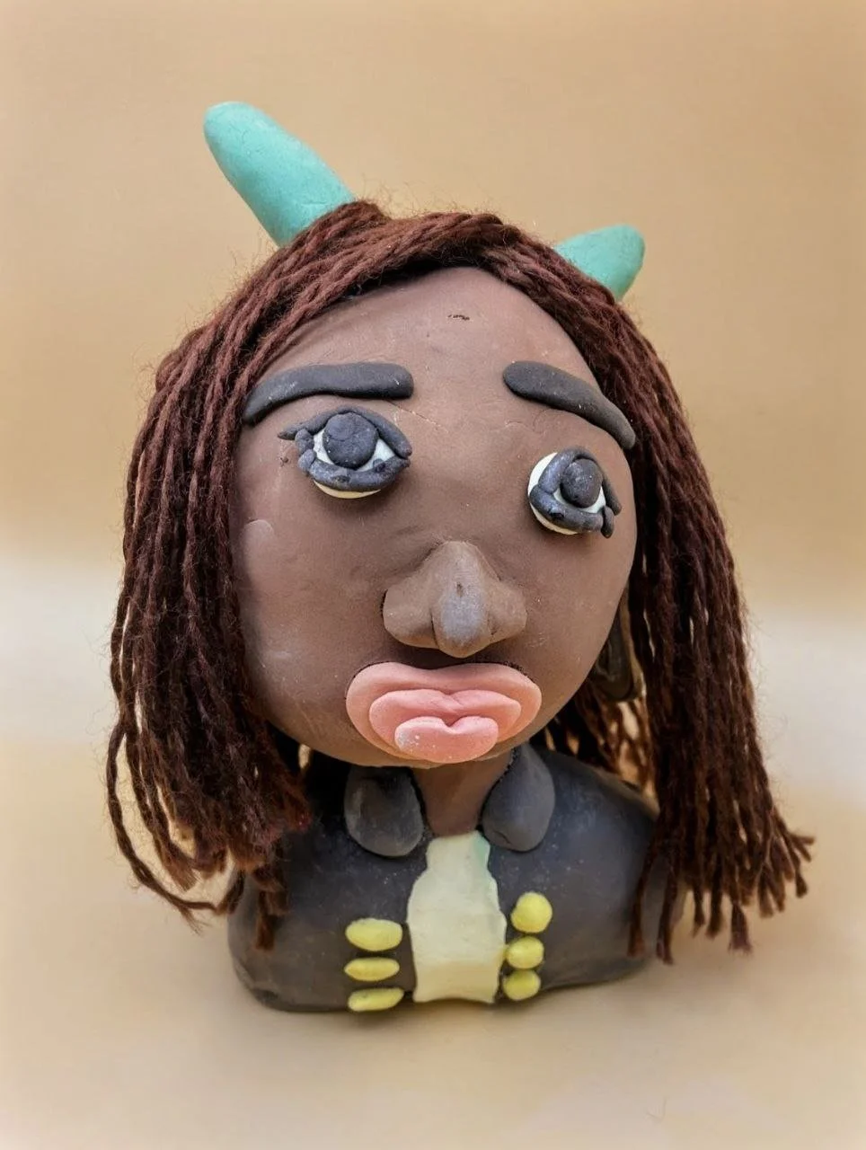 A clay sculpture of a woman with dark skin, brown hair styled in dreadlocks, wearing a black jacket with yellow buttons, and two light green horns on her head.