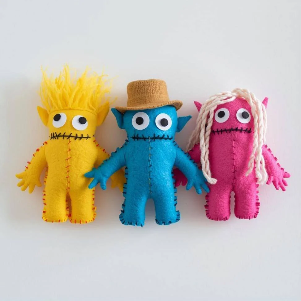 Three colorful plush dolls resembling monsters with large googly eyes, stitched mouths, and fabric features. The yellow doll has spiky hair, the blue doll wears a brown hat, and the pink doll has white yarn hair.